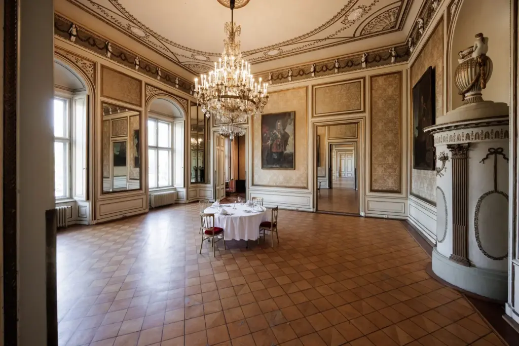 Ein prächtiger, verzierter Raum im Palais Auersperg mit hohen Decken, großen Fenstern, Kronleuchtern und eleganten Wanddekorationen. Ein runder Tisch mit vier Stühlen steht in der Mitte des großen, gefliesten Bodens.