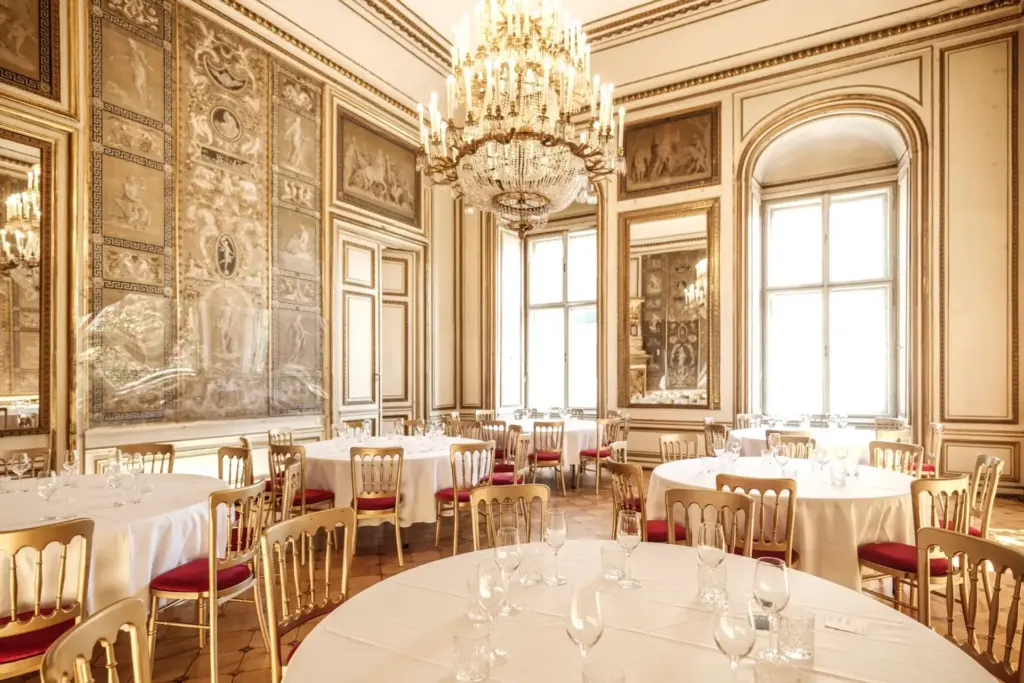 Eleganter Speisesaal im Palais Auersperg mit Kronleuchtern, goldverzierten Wänden, großen Spiegeln und mehreren runden Tischen mit weißen Tischtüchern und Gläsern, die von hohen Fenstern mit Tageslicht beleuchtet werden.