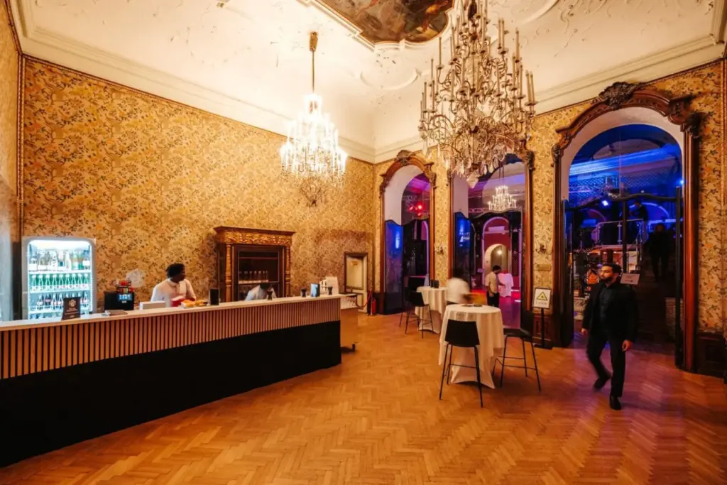 Eleganter Raum im Palais Auersperg mit verschnörkelten Kronleuchtern, goldgemusterten Tapeten und hohen Decken. Auf der linken Seite befindet sich ein Bartresen mit Personal, während Tische mit weißen Tüchern und ein paar Personen den stilvollen Bereich einnehmen.