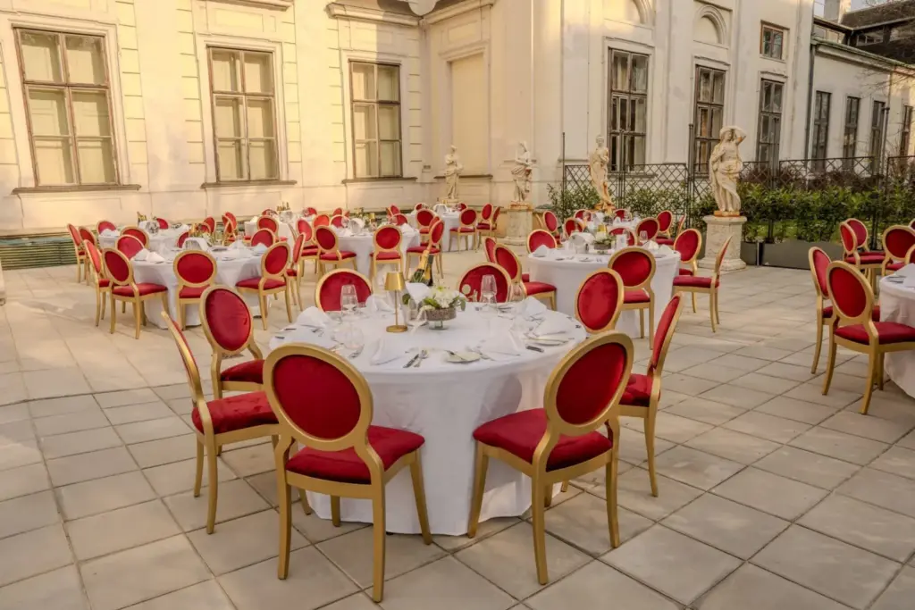 Elegantes Essen im Freien mit runden, weiß gedeckten Tischen, Tellern und Gläsern, umgeben von roten und goldenen Stühlen im Innenhof des Palais Auersperg, der mit klassischen Statuen und historischen Gebäuden geschmückt ist.