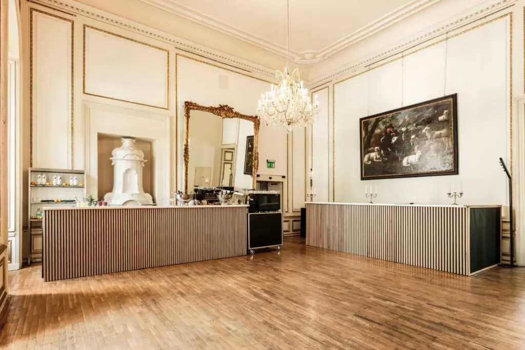 Eleganter, geräumiger Raum mit hohen Decken, Holzböden und einem großen Kronleuchter im Palais Auersperg. Verschnörkelte Wandleisten und eine Spiegelwand betonen einen minimalistischen Barbereich mit modernen Holztheken, Kunst und Getränkeregalen.