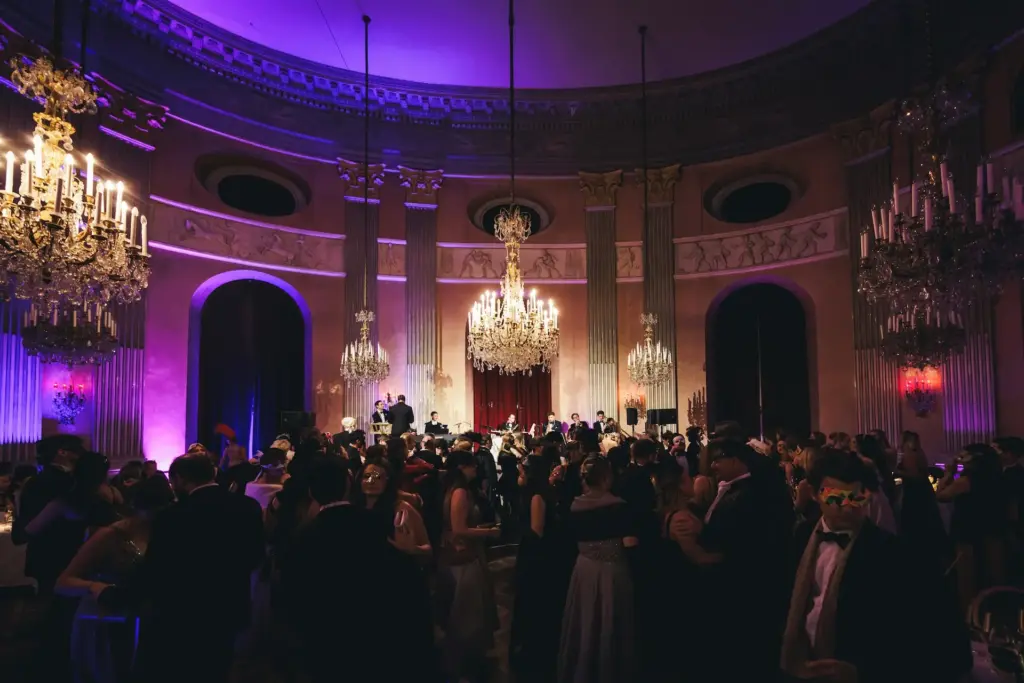 Ein großer, eleganter Ballsaal im Palais Auersperg mit Kronleuchtern und hohen Säulen ist gefüllt mit förmlich gekleideten Menschen, die Masken tragen, um einen Maskenball bei violetter Beleuchtung zu besuchen.