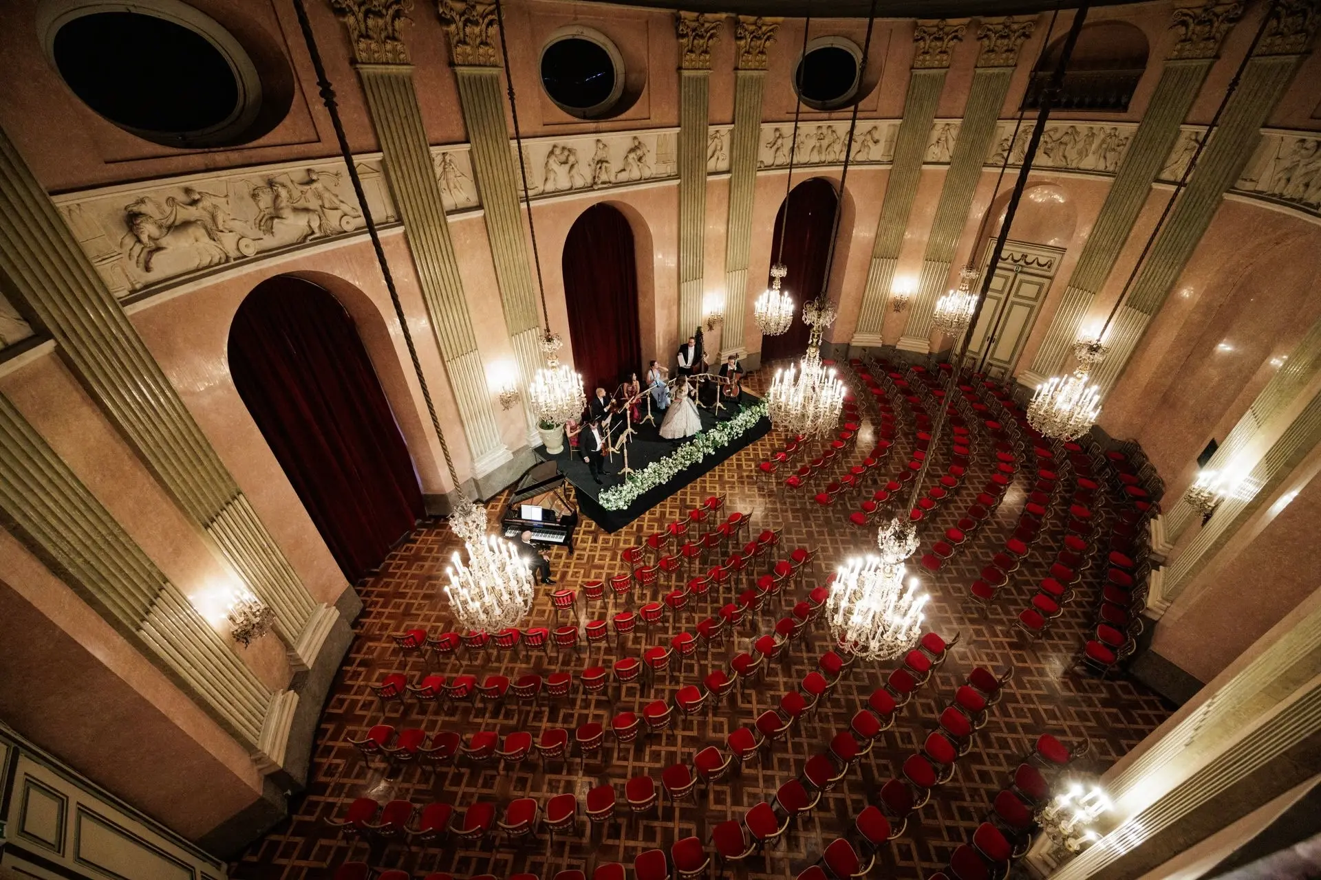 Ein großer, verzierter Saal mit Kronleuchtern und rot gepolsterten Stühlen, die in Bögen vor einer Bühne angeordnet sind, auf der Musiker auftreten. Der elegante Raum ist mit hohen Säulen, großen Bogentüren und dekorativen Wandreliefs ausgestattet.