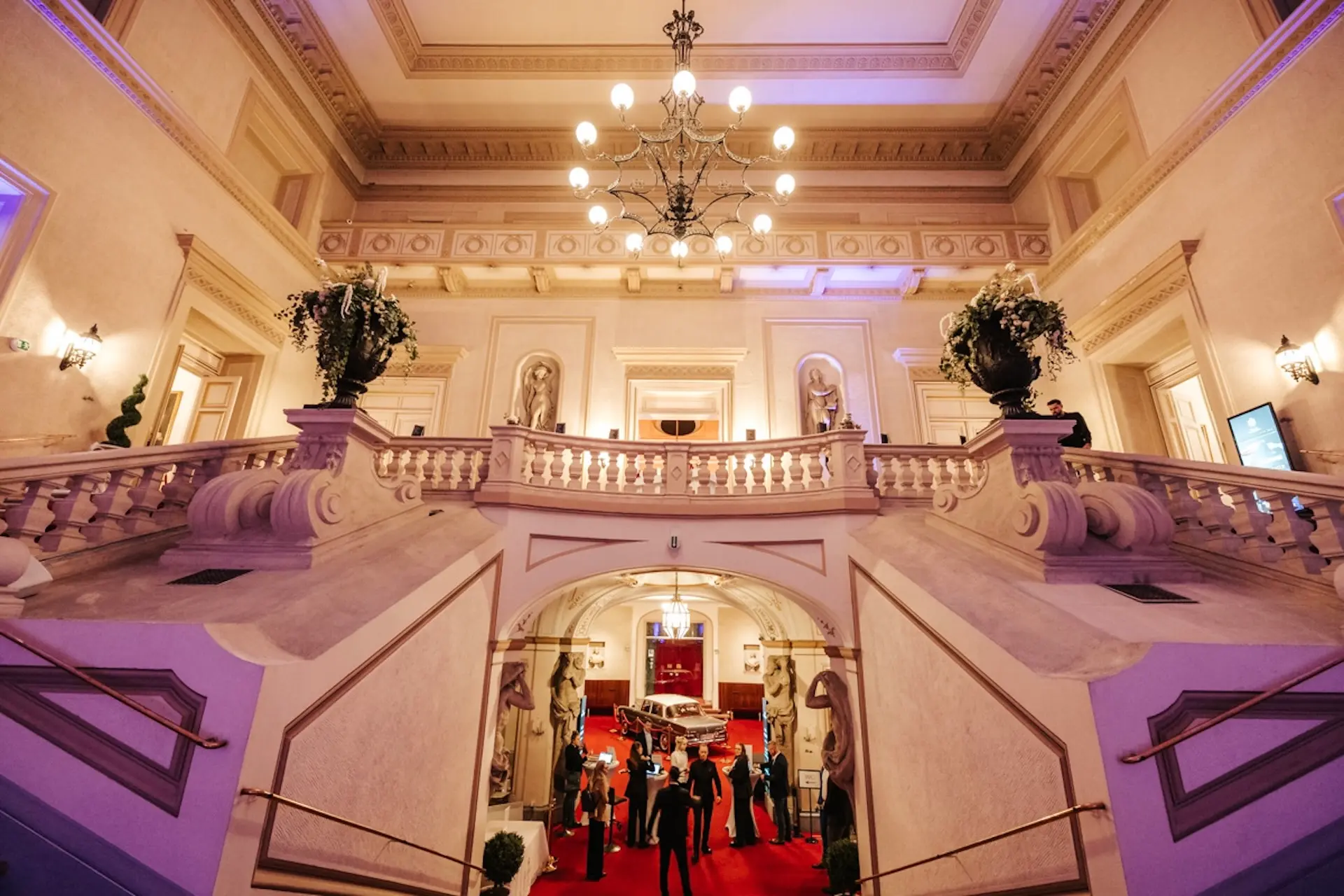 Große Treppe mit kunstvollem Geländer, Kronleuchtern, Statuen und Topfpflanzen in einer eleganten Halle. Mehrere formell gekleidete Personen stehen unten auf einem roten Teppich, was auf eine formelle Veranstaltung in einem historischen Gebäude hindeutet.