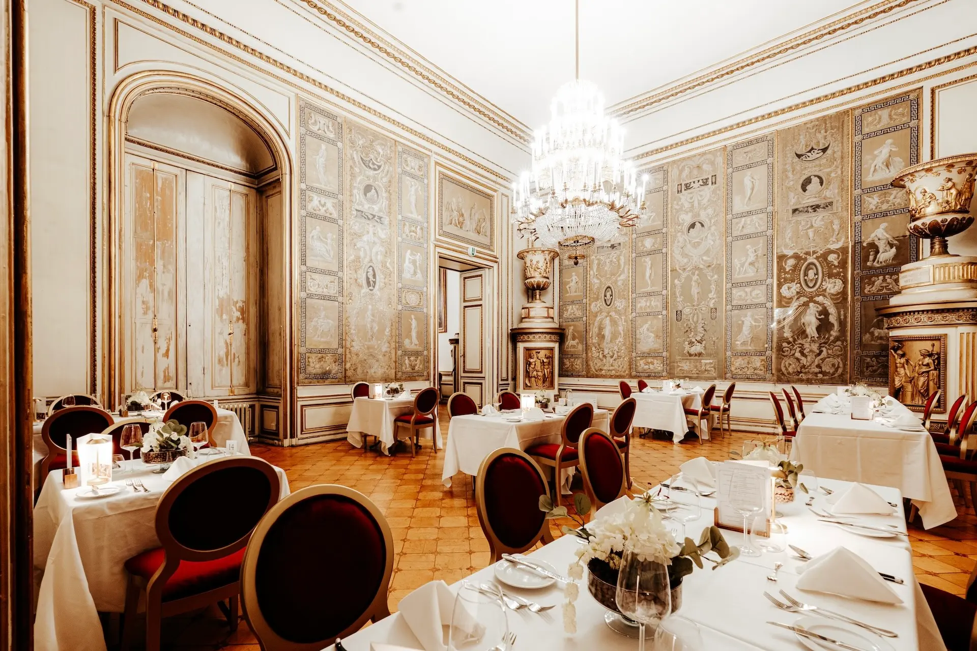 Eleganter Speisesaal mit verschnörkelten, goldverzierten Wänden, hoher Decke, großem Kronleuchter, runden Tischen mit weißer Bettwäsche, rot gepolsterten Stühlen und aufwändigem klassischen Dekor. Die Atmosphäre ist luxuriös und formell.