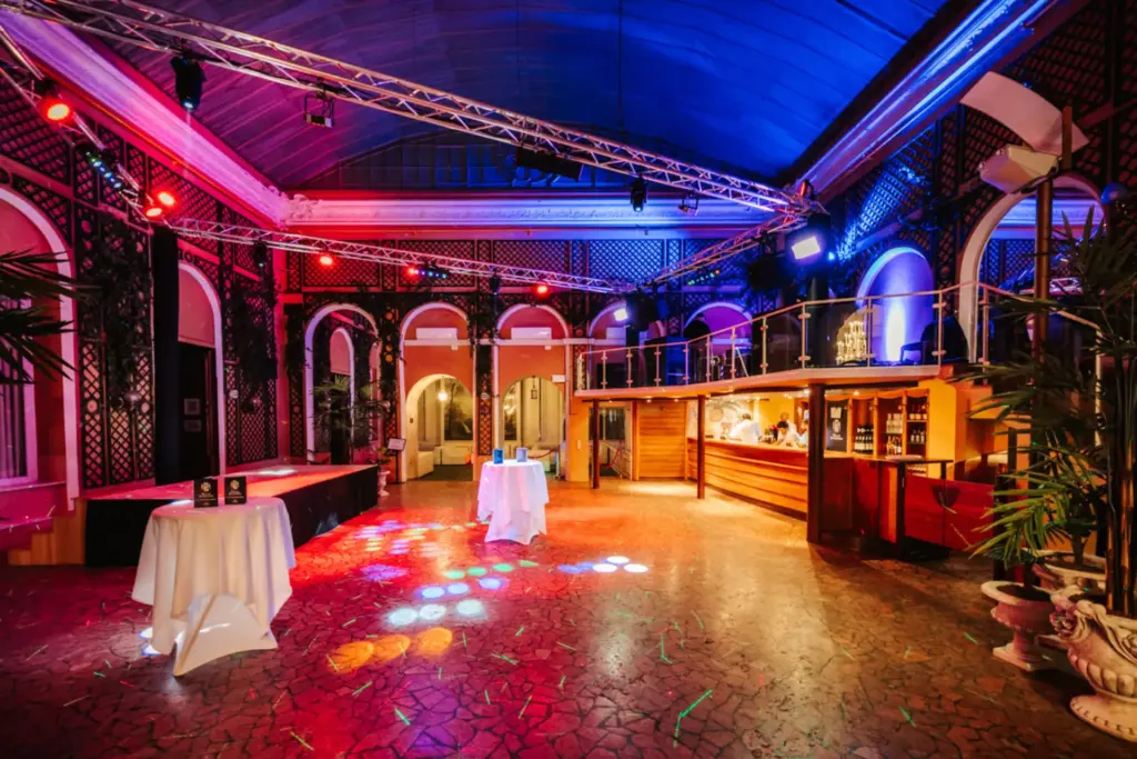 Ein geräumiger, eleganter Veranstaltungssaal im Palais Auersperg mit farbiger Beleuchtung, leeren Tischen mit weißen Tüchern, einem Barbereich und gewölbten Fenstern. Die hohe, geschwungene Decke und der gemusterte Boden machen den Raum bereit für die Gäste.