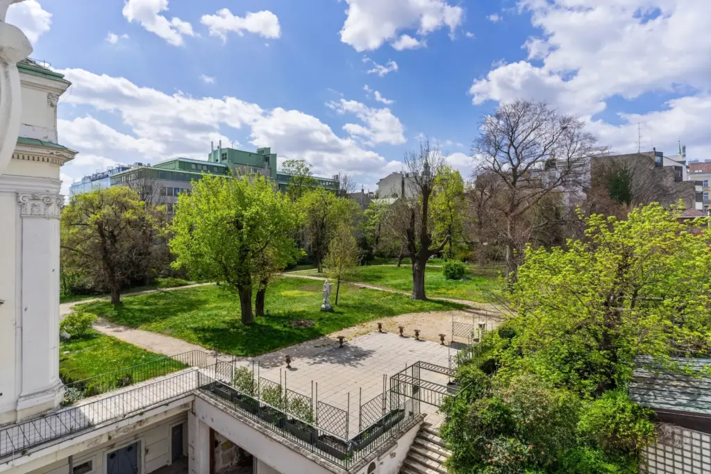 Ein sonniger Stadtpark mit grünen Rasenflächen, Bäumen und Spazierwegen, der von Gebäuden begrenzt wird. Im Vordergrund befindet sich eine gepflasterte Terrasse mit einer Treppe, die hinunter in den Gartenbereich führt. Der Himmel ist hell mit vereinzelten Wolken.