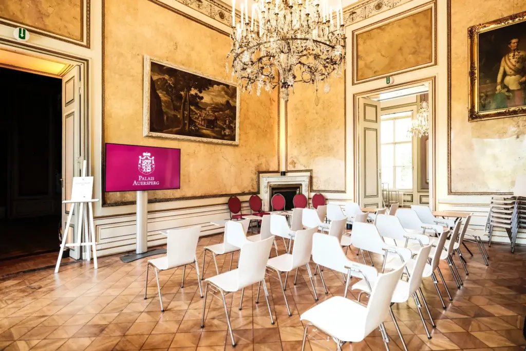 Eleganter Raum mit verschnörkelten Kronleuchtern, großen Gemälden und hohen Fenstern. Weiße Stühle sind in Reihen vor einem Bildschirm angeordnet, auf dem das Palais Albrizzi zu sehen ist. Der Raum ist klassisch eingerichtet und verfügt über einen Kamin.
