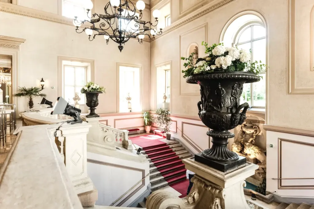 Elegante Innentreppe mit verschnörkeltem Geländer, großen Fenstern und dekorativen schwarzen Urnen mit weißen Blumen. Ein roter Teppich führt die Treppe hinunter, und ein großer Kronleuchter hängt von der hohen Decke herab.