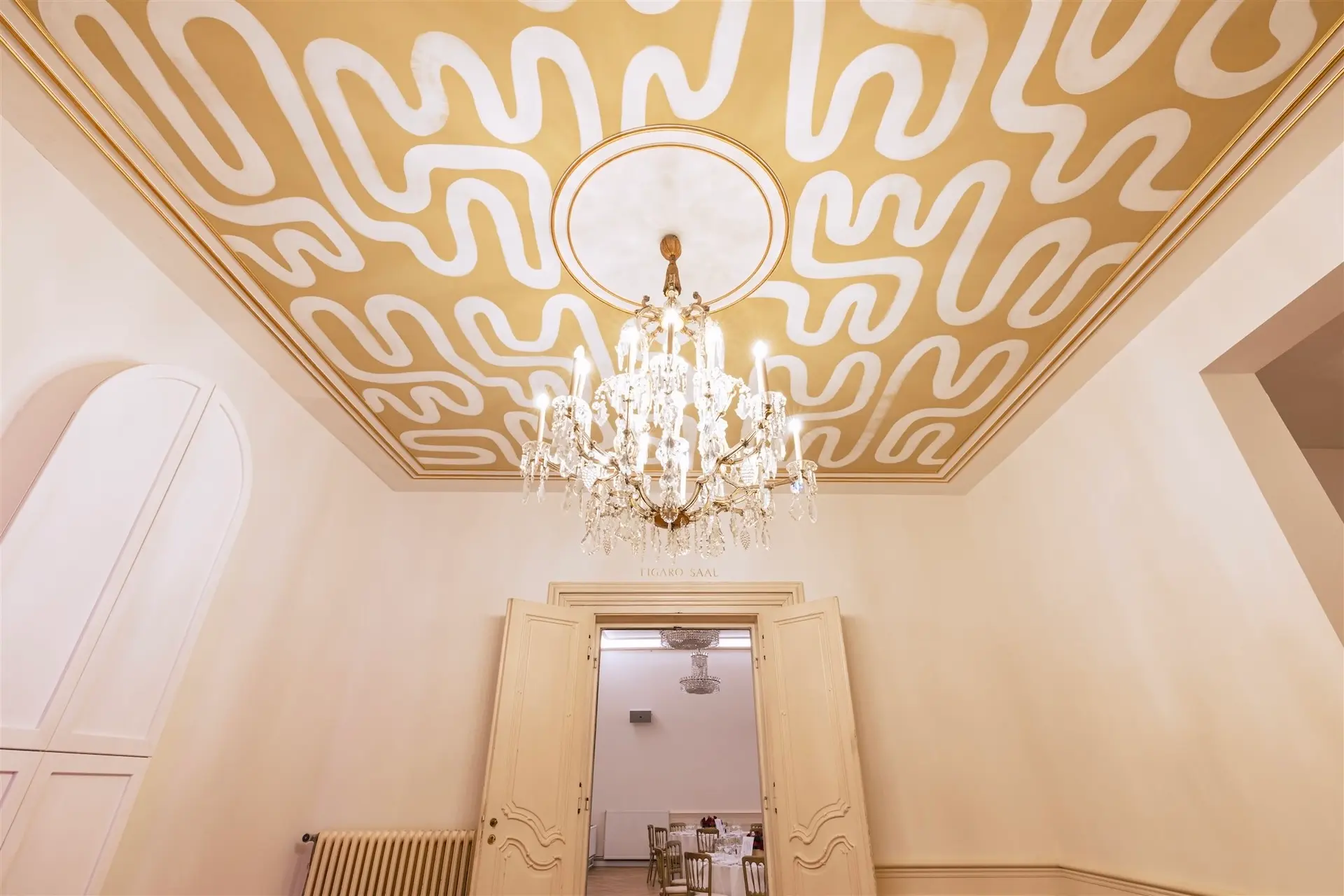 In einem eleganten Raum mit hellen Wänden und Flügeltüren hängt ein verschnörkelter Kristalllüster von einer mit kühnen, abstrakten Mustern in Gold und Weiß verzierten Decke herab.