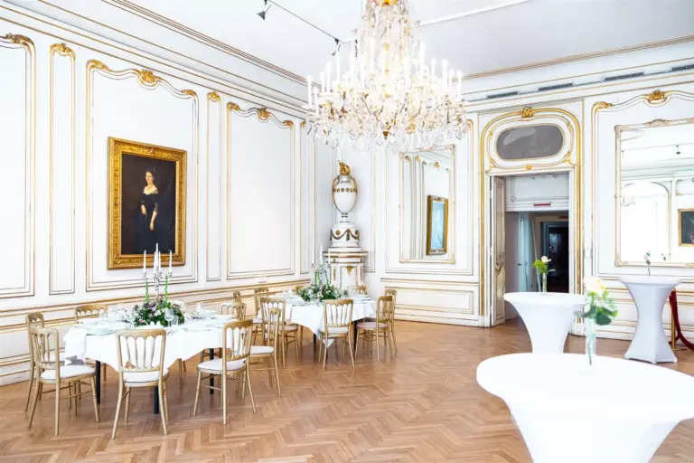 Eleganter Bankettsaal mit weißen Wänden, goldenen Verzierungen, einem großen Kronleuchter und Parkettböden. Runde Tische mit weißen Tischtüchern und Blumenkränzen sind für eine Veranstaltung gedeckt. Ein großes Porträt hängt an der Wand.