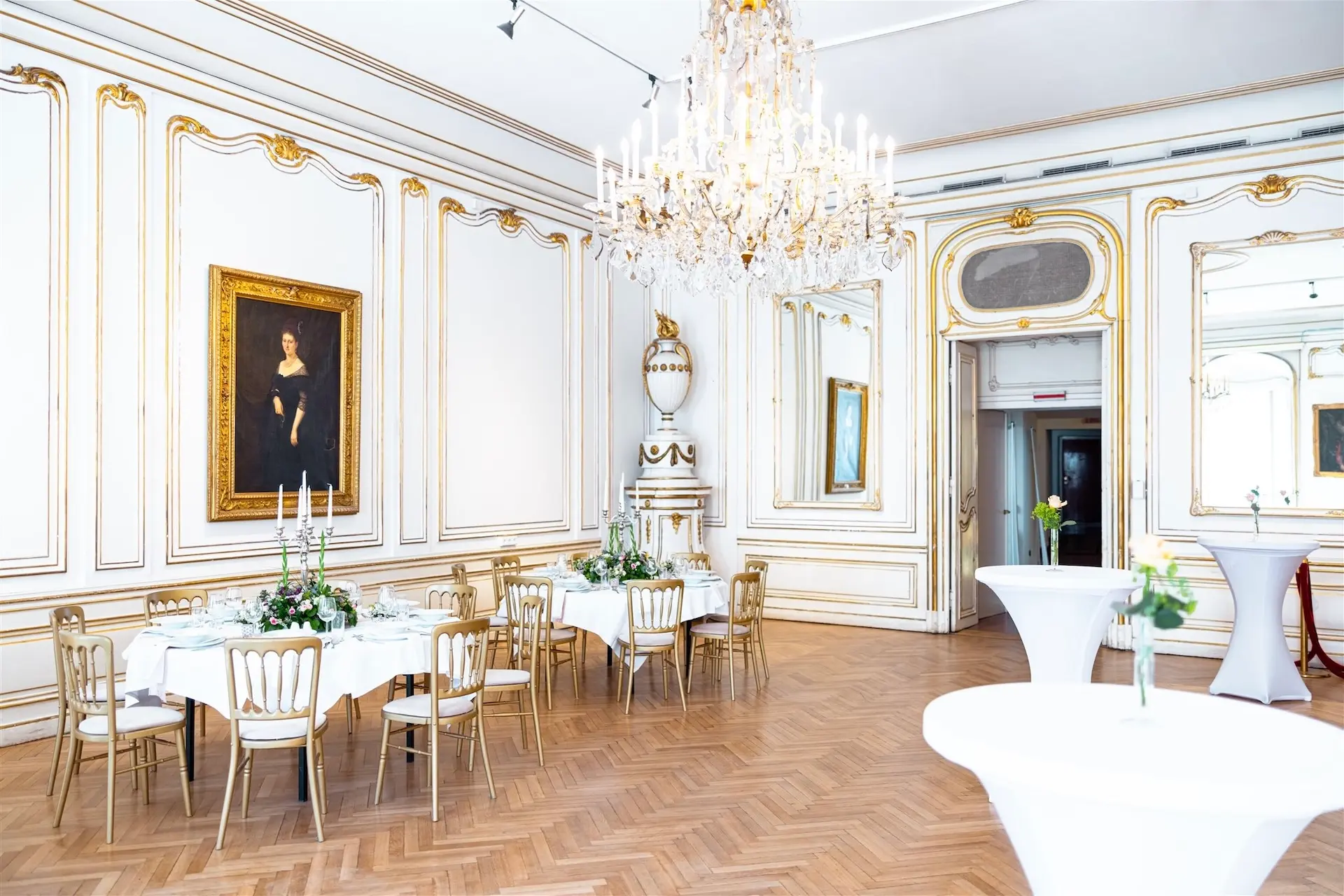 Eleganter Bankettsaal mit weißen Wänden, goldenen Verzierungen, einem großen Kronleuchter und Parkettböden. Runde Tische mit weißen Tischtüchern und Blumenkränzen sind für eine Veranstaltung gedeckt. Ein großes Porträt hängt an der Wand.