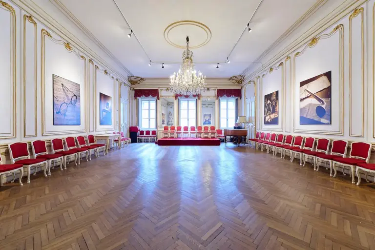 Eleganter Ballsaal mit Fischgrätenholzboden, roten und goldenen Stühlen an den Wänden, einem großen Kronleuchter, großen Fenstern mit roten Vorhängen und gerahmten Kunstwerken an weißen Wänden mit Goldverzierung.