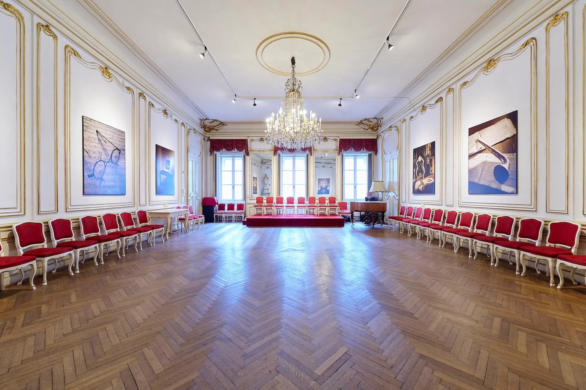 Eleganter Ballsaal mit Fischgrätenholzboden, roten und goldenen Stühlen an den Wänden, einem großen Kronleuchter, großen Fenstern mit roten Vorhängen und gerahmten Kunstwerken an weißen Wänden mit Goldverzierung.