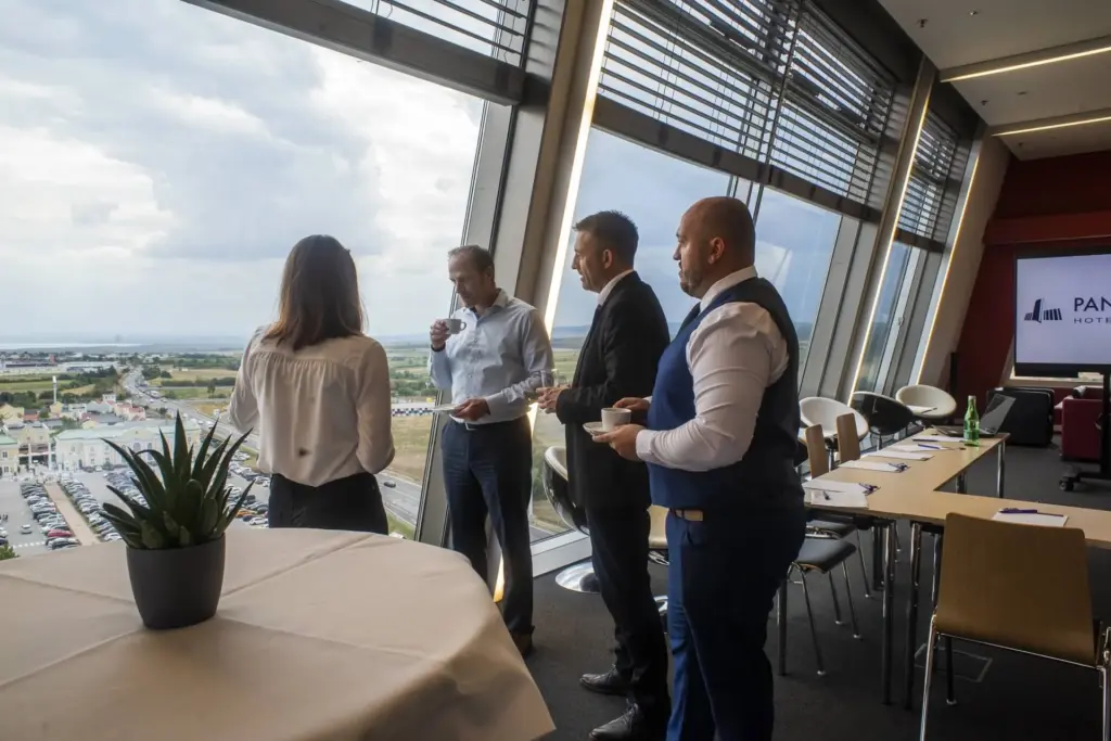 Vier Personen in Geschäftskleidung stehen an großen Fenstern, unterhalten sich und halten Getränke in einem modernen Konferenzraum mit Tischen und Stühlen und blicken auf eine Stadtlandschaft.