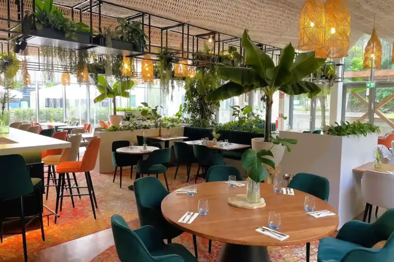 Ein modernes Restaurant mit Grünpflanzen, Hängelampen, runden Holztischen mit Geschirr und Gläsern, Samtstühlen in Teal und Orange und großen Fenstern, die natürliches Licht hereinlassen.