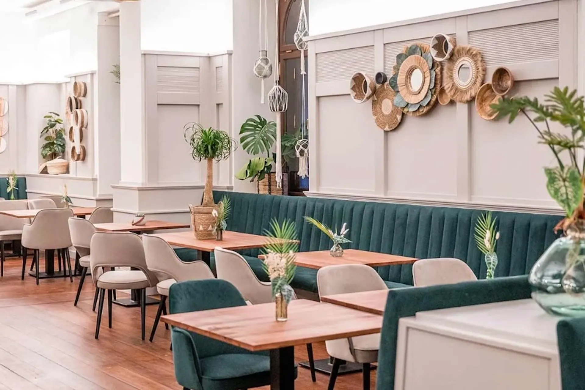 Ein stilvolles Restaurant mit Sitzgelegenheiten aus grünem Samt, Holztischen, beigen Stühlen, Topfpflanzen, Holzböden und dekorativen Weidenkörben an einer hellen Wand.