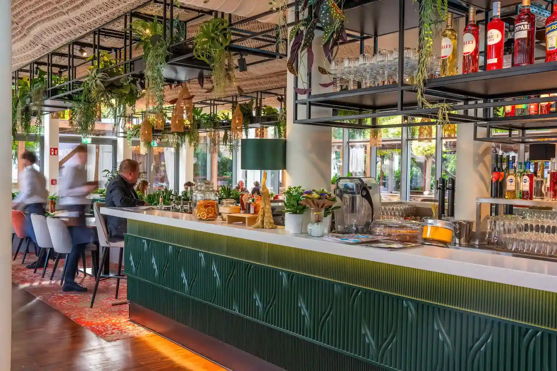 Eine moderne Bar mit einem grün gefliesten Tresen, hängenden Pflanzen, Flaschen und Glaswaren. Die Leute sitzen an Tischen in der Nähe, und große Fenster lassen natürliches Licht herein und schaffen eine lebendige und einladende Atmosphäre.
