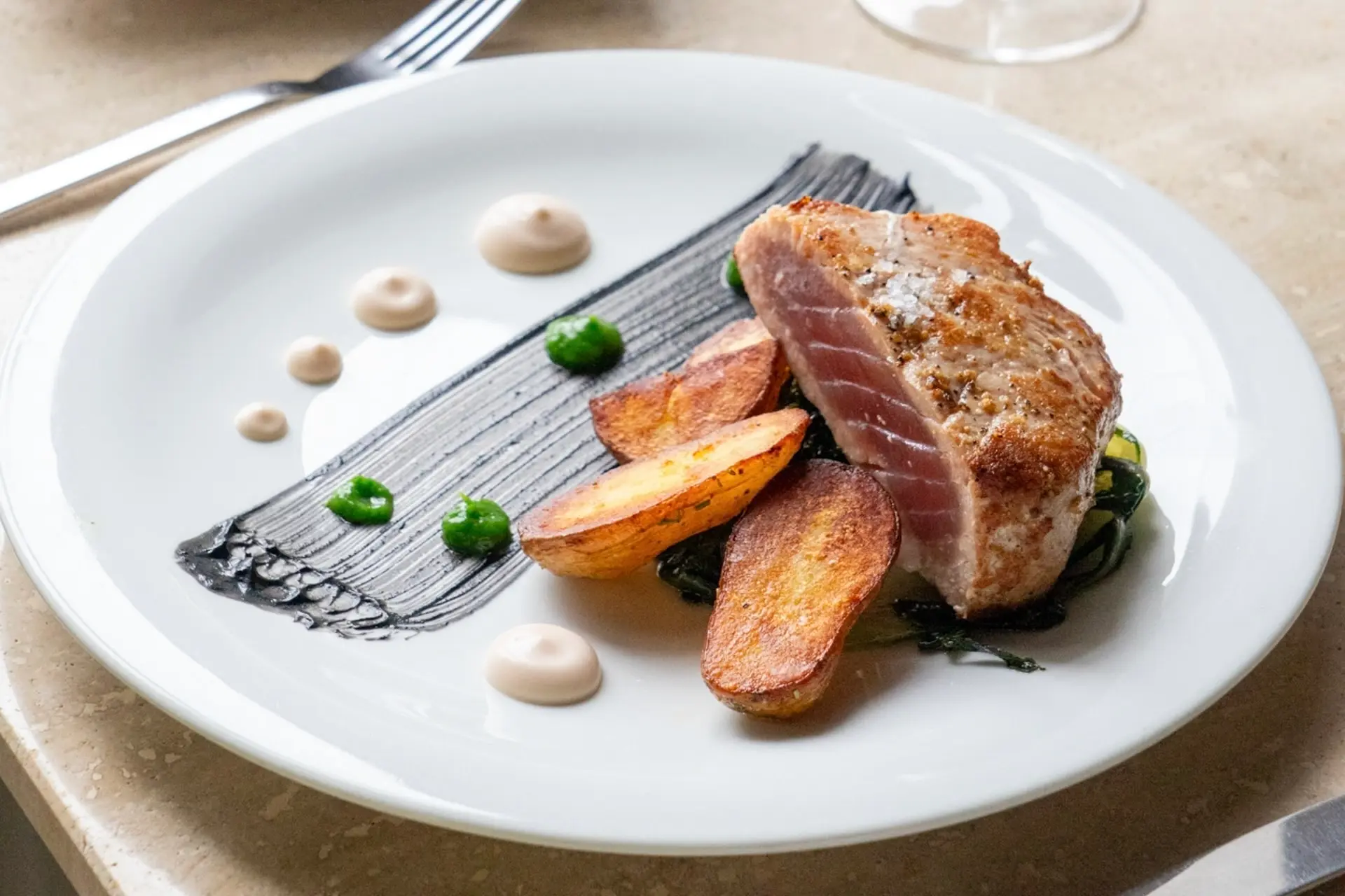 Ein wunderschön angerichtetes Gericht mit einem gebratenen Thunfischsteak, gerösteten Kartoffelecken, grünen Soßenpunkten, beigen Soßentupfern und einem Pinselstrich schwarzer Soße auf einem weißen Teller, mit einer Gabel im Hintergrund.