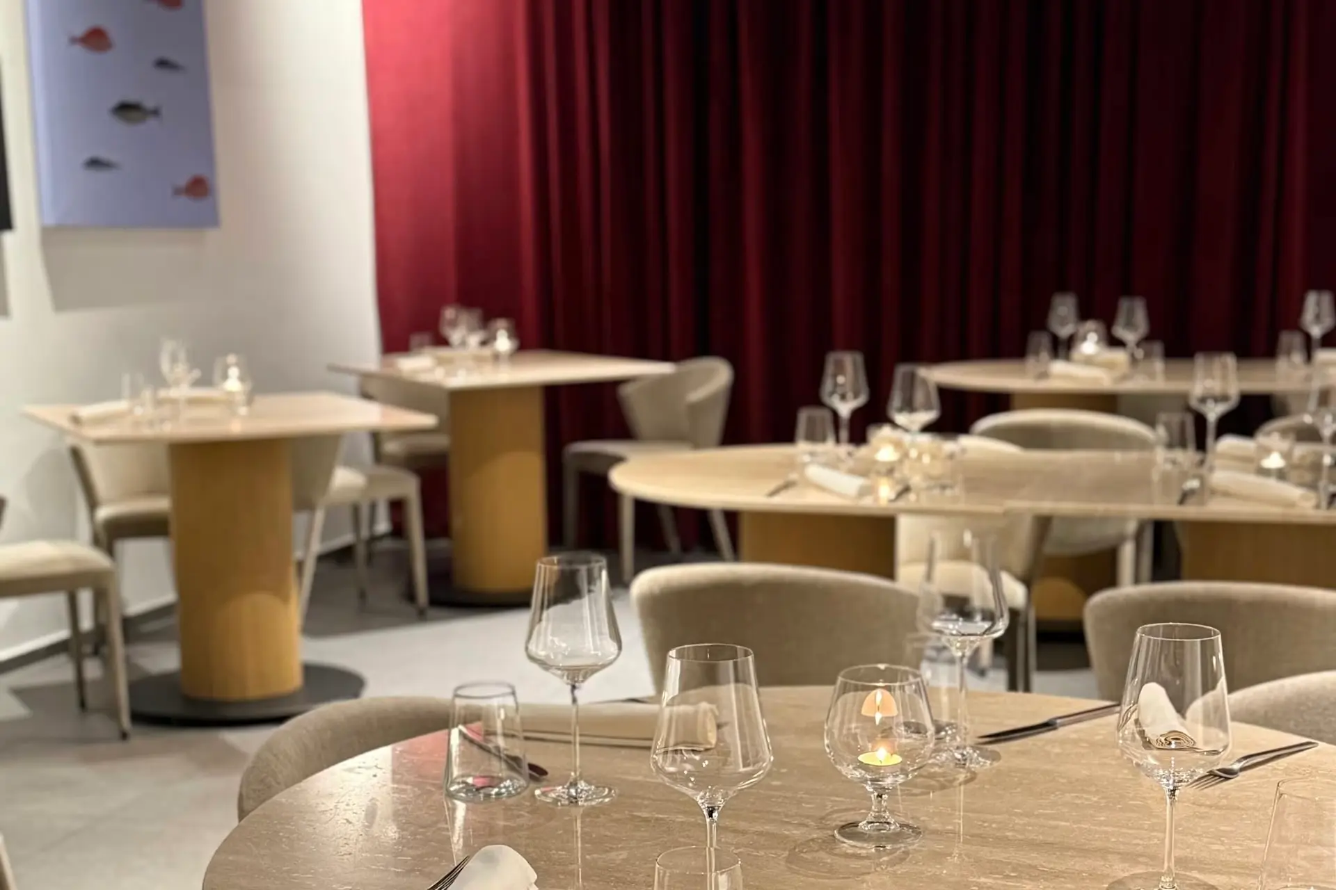 Ein moderner Restaurantbereich mit runden Tischen, beigen Stühlen, klaren Weingläsern und gefalteten Servietten. Rote Vorhänge bedecken eine Wand, und an der weißen Wand im Hintergrund hängt ein Gemälde mit kleinen Fischen.