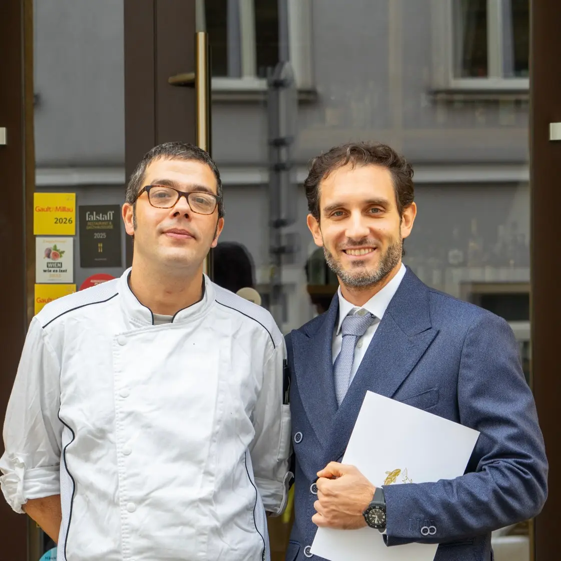 Zwei Männer stehen nebeneinander; der eine trägt eine weiße Kochuniform und eine Brille, der andere einen blauen Anzug, eine Krawatte und hält ein Dokument in der Hand. Sie stehen vor einem Gebäude mit Glastüren und Preisschildern.