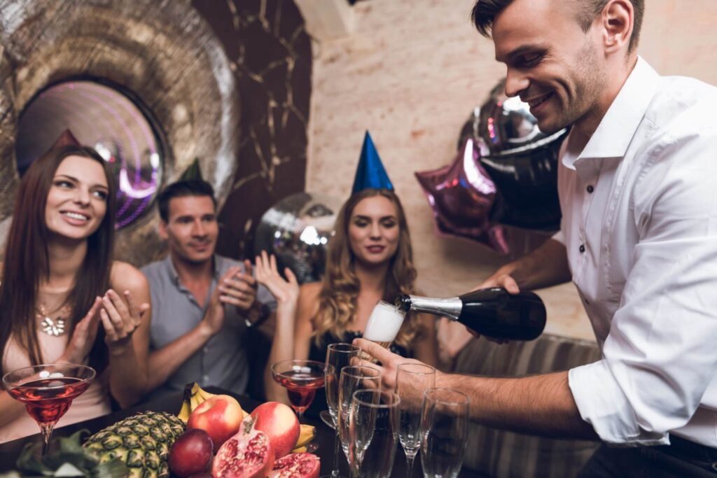 Ein Mann schenkt bei einer lebhaften Party in einem Partykeller in Wien Sekt in Gläser ein, während drei Personen mit Partyhüten lächeln und klatschen. Festliche Dekoration und ein Tisch mit Obst und Getränken tragen zur fröhlichen Atmosphäre bei.