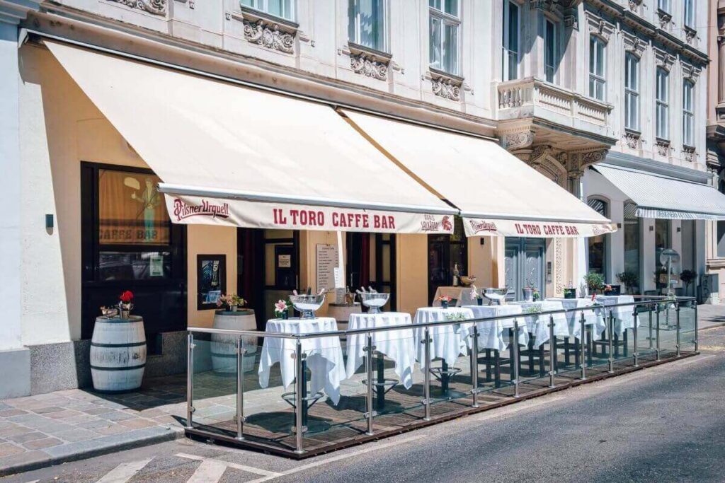 Der Außensitzbereich der Il Toro Caffe Bar mit mehreren Tischen, die mit weißen Tüchern bedeckt und mit Gläsern und Blumen geschmückt sind - ein einladender Ort neben einem verzierten historischen Gebäude, perfekt, wenn Sie eine Partylocation in der Stadt mieten möchten.