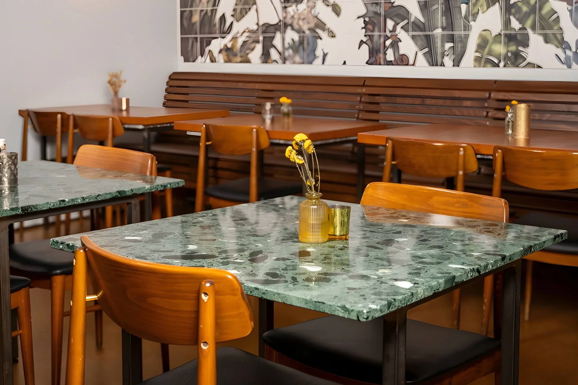 Ein gemütlicher Café-Essbereich mit Tischen aus grünem Marmor, Holzstühlen mit schwarzen Sitzen, kleinen Vasen mit gelben Blumen und einer Holzbank an der Wand mit einem Wandgemälde aus Blättern darüber.