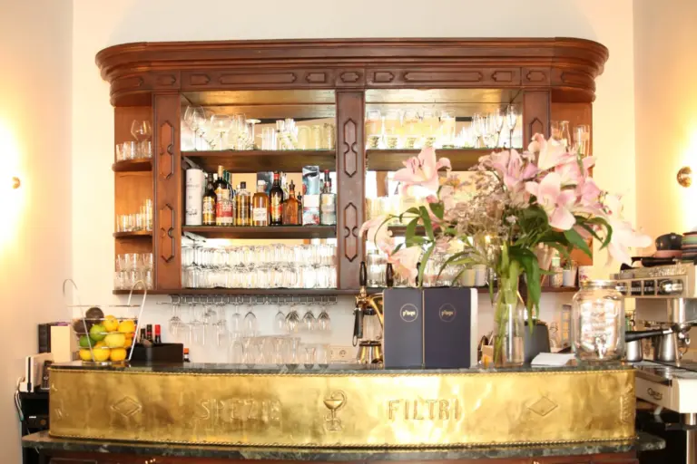 Eine polierte Bar mit einer Messingtheke, Holzregale mit Flaschen und Gläsern, ein großer Strauß rosa Lilien und eine Kaffeemaschine schaffen eine warme und einladende Atmosphäre.