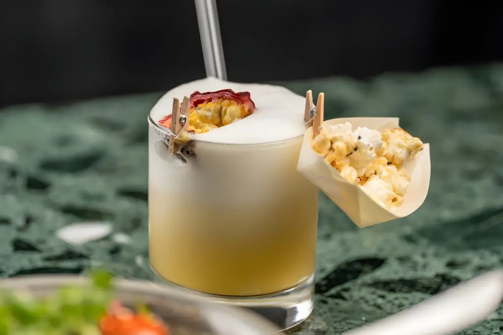 Ein Cocktail mit weißer Schaumkrone, garniert mit einer Scheibe Trockenobst und einer abgeschnittenen Passionsfrucht, serviert in einem Glas mit einem Metallstrohhalm und einem kleinen Papierkegel mit Popcorn am Rand.