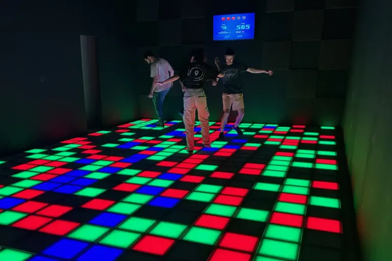 Pixel Arena Salzburg Drei Personen spielen in einem dunklen Raum auf einem beleuchteten Boden mit bunten Fliesen in Rot, Grün und Blau. An der Wand im Hintergrund hängt eine digitale Anzeigetafel, die die Punkte anzeigt.