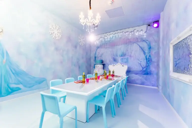 Ein pastellblauer und weißer Partyraum zum Thema Frozen mit einem langen Tisch, blauen Stühlen, Wandbildern mit eisigen Szenen, Schneeflocken-Dekoration, einem Kronleuchter und Partyzubehör auf dem Tisch.