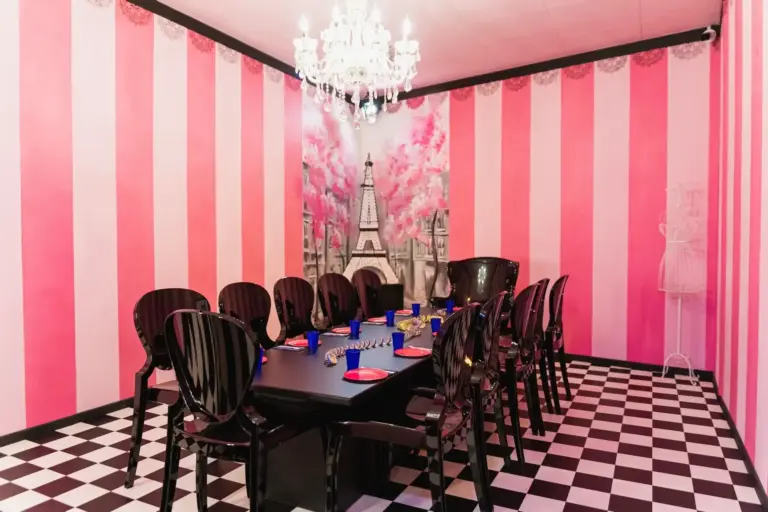 Ein Esszimmer mit rosa-weiß gestreiften Wänden, einem Wandgemälde des Eiffelturms, einem Kronleuchter, schwarzen Stühlen um einen langen schwarzen Tisch mit roten Tellern und blauen Tassen und einem schwarz-weiß karierten Boden.
