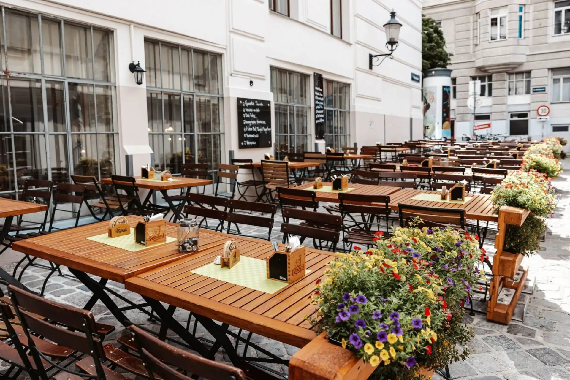 Außencafé mit Reihen von Holztischen und -stühlen auf einem gepflasterten Innenhof, der mit bunten Blumenkübeln geschmückt ist. Das Café befindet sich neben einem weißen Gebäude mit großen Fenstern und einer Speisekarte auf einer Kreidetafel.