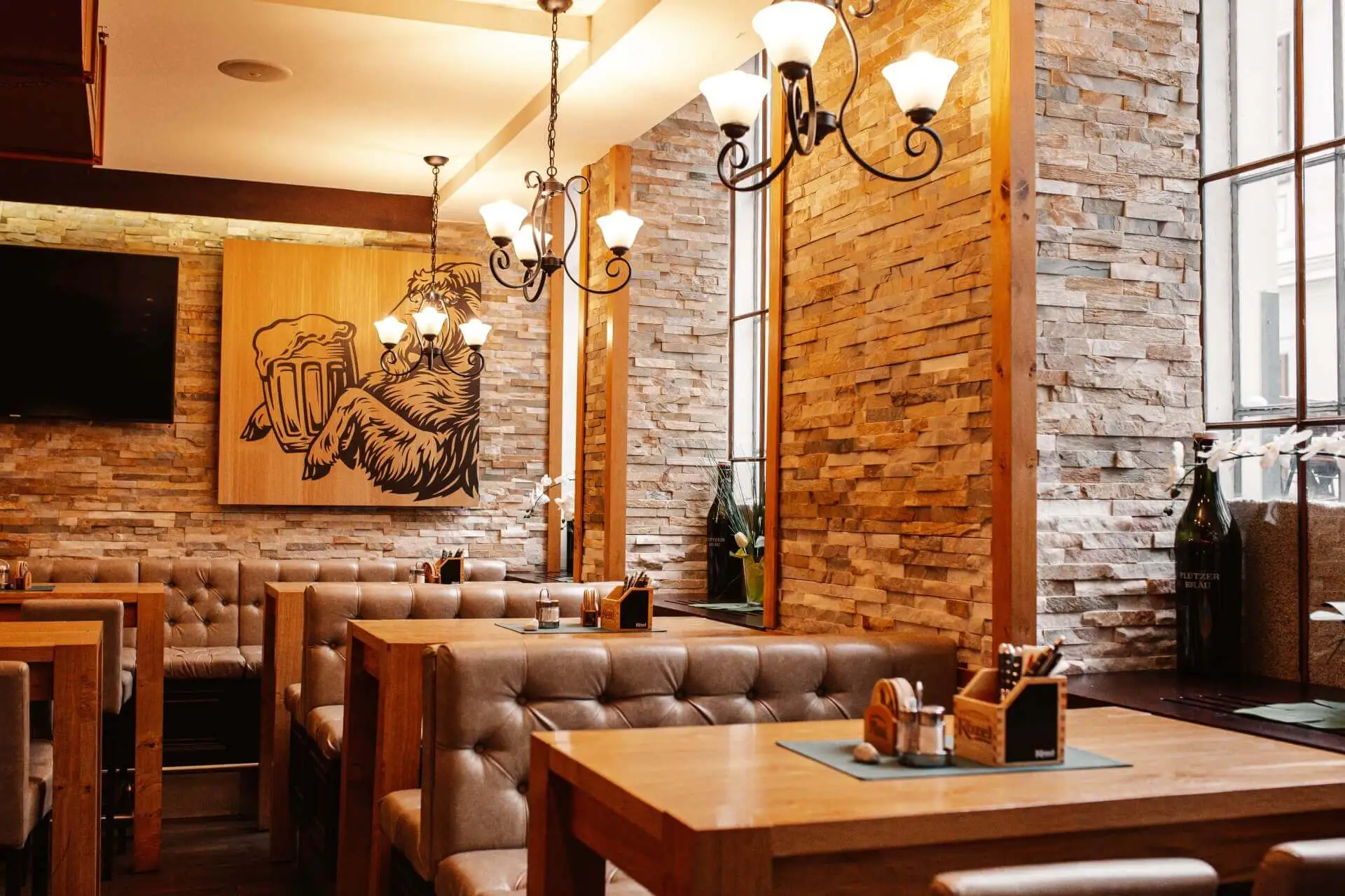 Ein gemütliches Restaurant mit Steinwänden, Holztischen, beigefarbenen, getufteten Sitzmöbeln und eleganten Kronleuchtern. Ein großes Gemälde eines Faultiers, das Bierkrüge hält, ziert die Wand. Das Sonnenlicht strömt durch hohe Fenster herein.