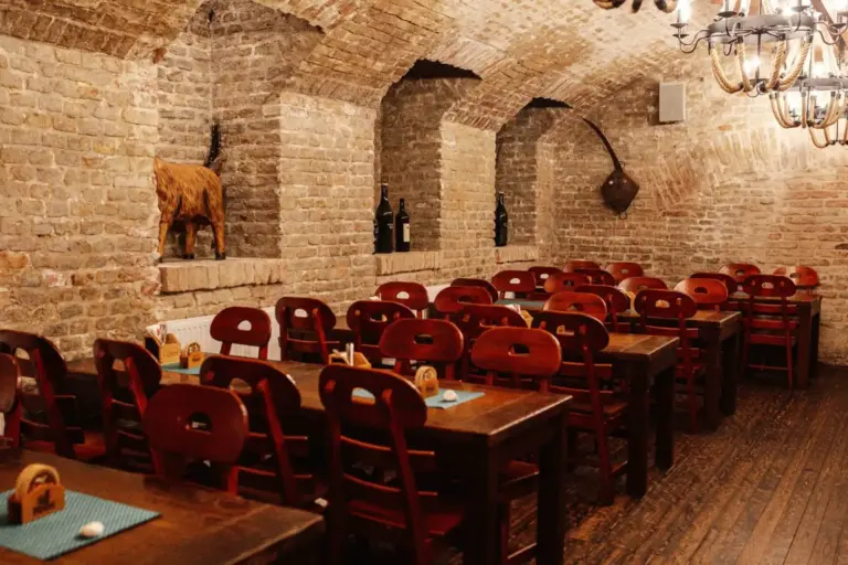 Ein rustikales Restaurant mit gewölbten Ziegelwänden und -decken, Reihen von Holztischen und -stühlen, Weinflaschen in Nischen und einer dekorativen Tierskulptur auf einem Steinsims.