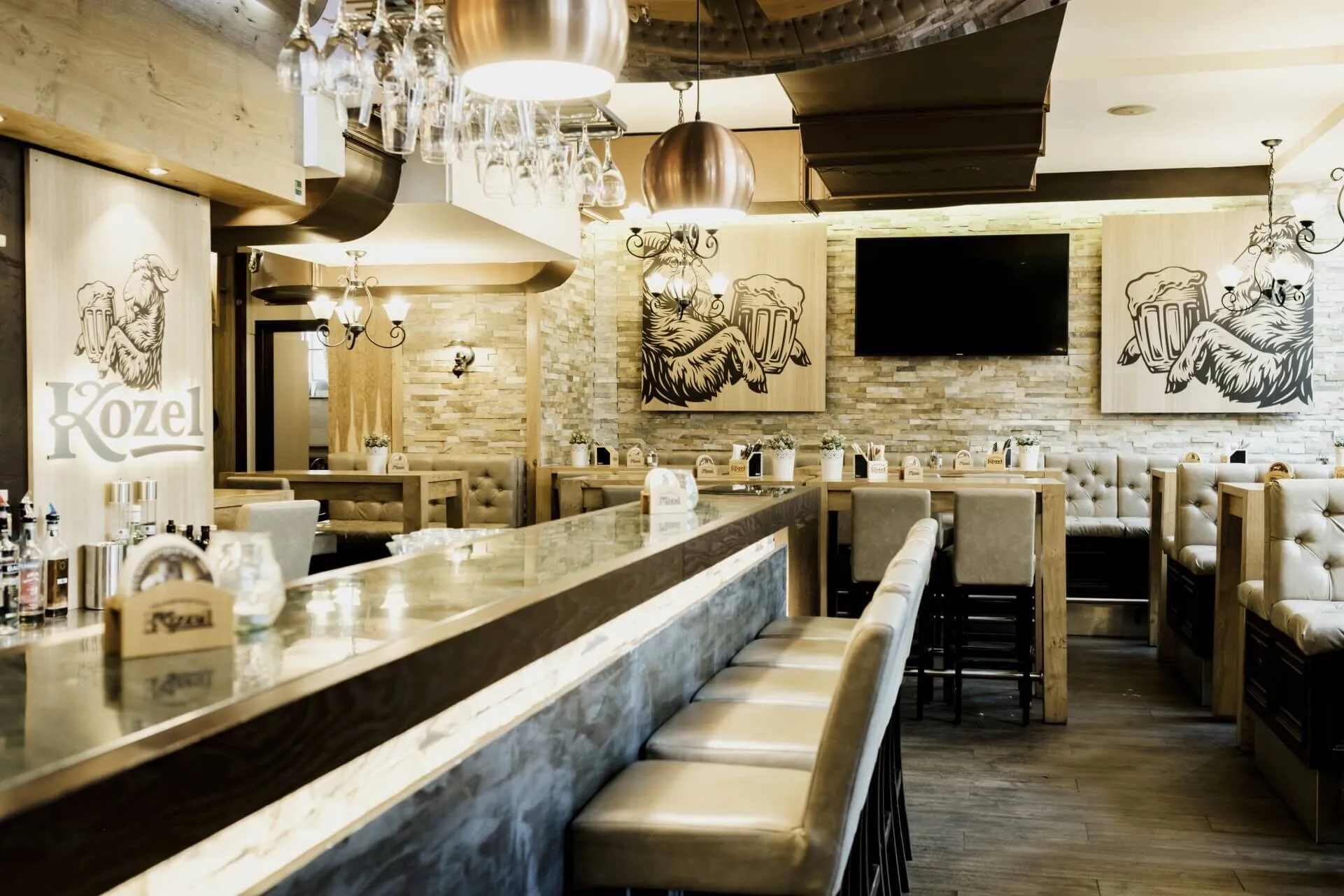 Eine moderne Bar mit einem langen Tresen, hohen Hockern, Glasregalen über dem Kopf und gedämpfter Beleuchtung. Gemütliche Sitzecken und Kunstwerke, die Ziegen mit Bierkrügen zeigen, schmücken die Wände; über einer Sitzecke ist ein großer Fernseher angebracht.