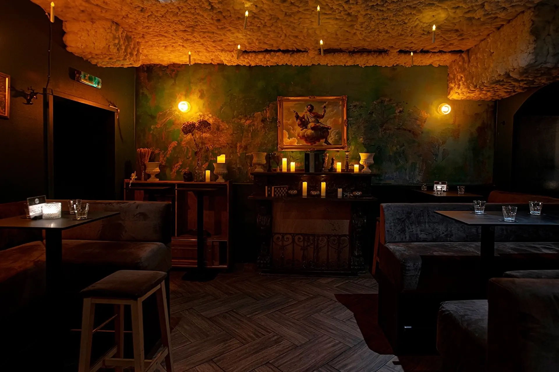 Eine schwach beleuchtete Bar mit bequemen Sitzgelegenheiten, einer strukturierten Decke, leuchtenden Wandlampen, Kerzen und einem Gemälde über einem Kamin schaffen ein gemütliches, intimes Ambiente.