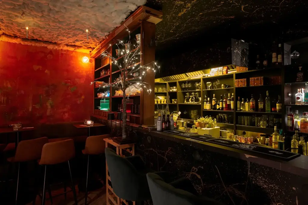 Eine schummrig beleuchtete, stilvolle Bar mit schwarzem Marmortresen, grünen Samtstühlen, Flaschenregalen, warmen roten Wänden und dekorativer Beleuchtung, die eine gemütliche und einladende Atmosphäre schafft.