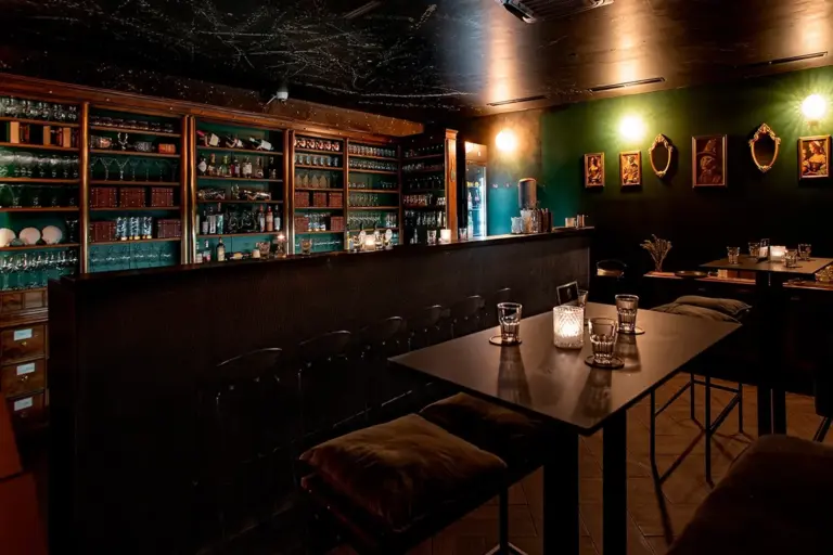 Eine schwach beleuchtete Bar mit einem dunklen Tresen, dahinter Regale mit Flaschen und Gläsern, Kerzen auf den Tischen, Barhocker mit Kissen und grüne Wände, die mit gerahmten Bildern geschmückt sind. Die Atmosphäre ist gemütlich und intim.