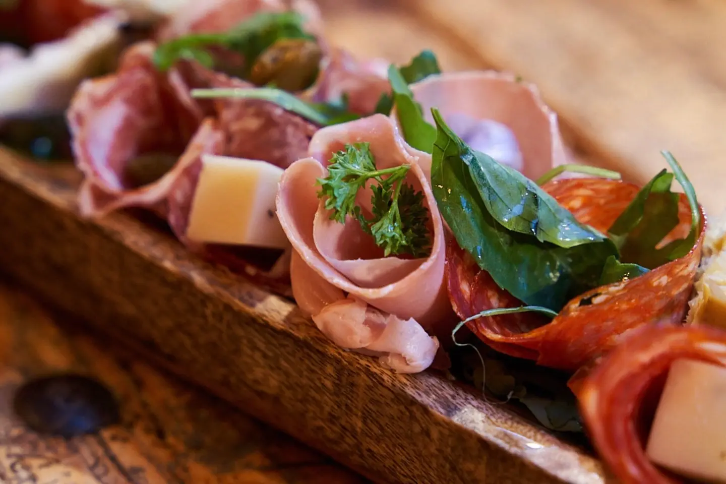 Nahaufnahme eines Holzbretts mit verschiedenen Wurstwaren, darunter gerollte Wurstwaren, Käsewürfel, frisches Grün und mit Kräutern garniert.