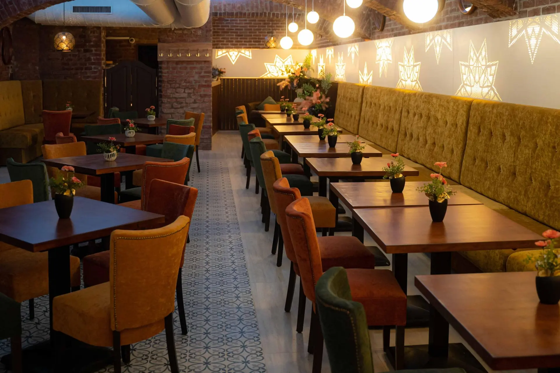 Gemütliches Restaurantinterieur mit Samtstühlen, Holztischen, kleinen Blumenvasen und gemusterten Bodenfliesen. Warme Beleuchtung und sternförmige Wandleuchten sorgen für eine einladende Atmosphäre. Sichtbare Backsteinwände und eine Gewölbedecke sind zu sehen.