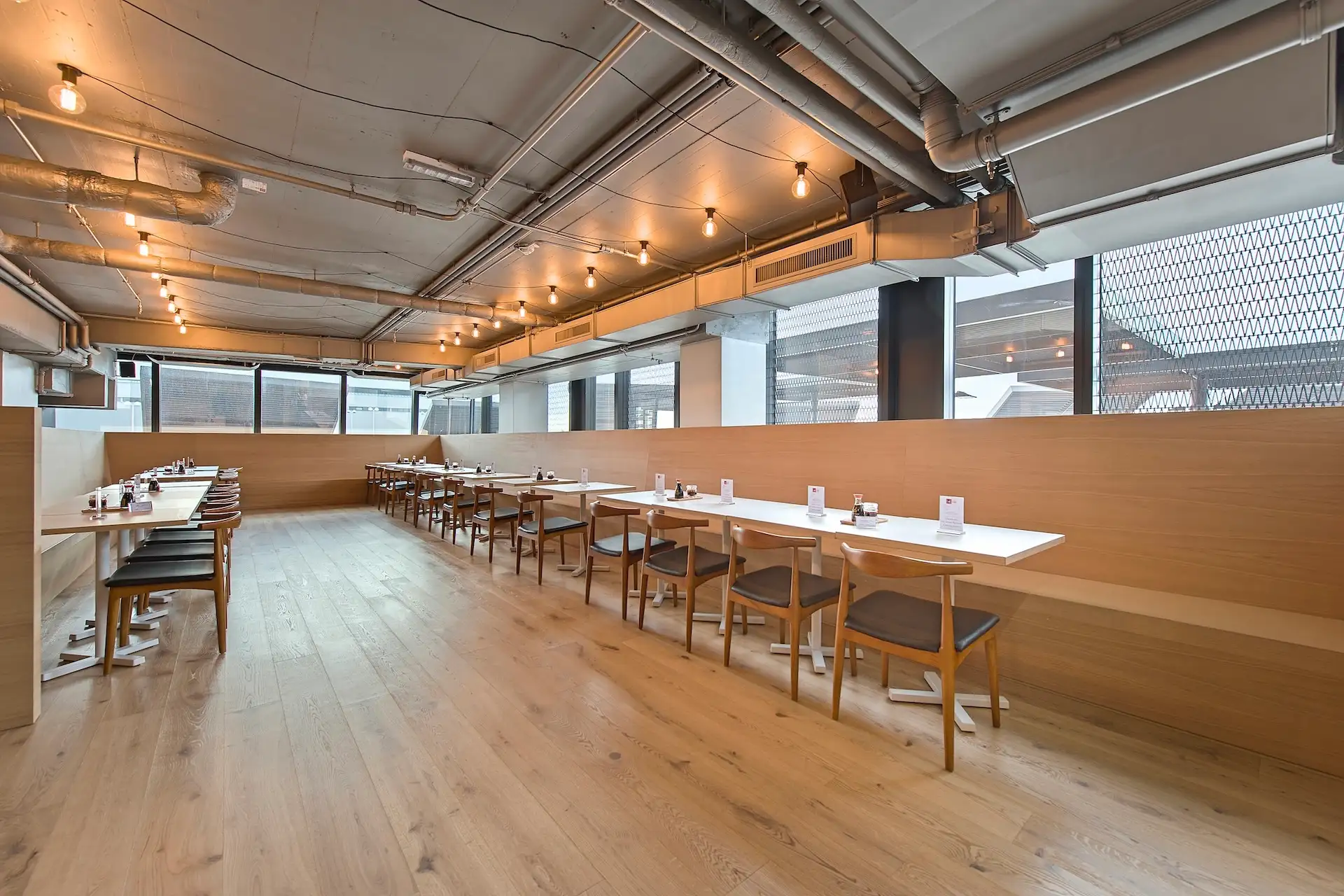 Ein modernes, geräumiges Restaurant mit Holzböden, Tischen und Stühlen aus hellem Holz, freiliegenden Rohrleitungen an der Decke und großen Fenstern, durch die Tageslicht hereinströmt. Die Tische sind ordentlich entlang der Wand angeordnet.