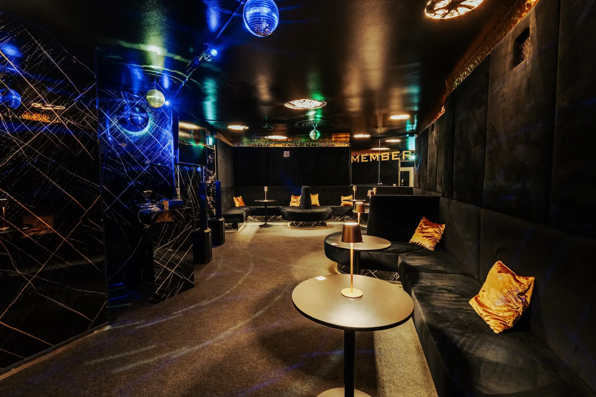 Eine schummrig beleuchtete Lounge mit schwarzen Wänden, runden Tischen, goldenen Kissen auf schwarzen Sofas und mehreren von der Decke hängenden Discokugeln. Die Atmosphäre ist modern und luxuriös, mit sanfter Beleuchtung und einem "MEMBER"-Schild im Hintergrund.