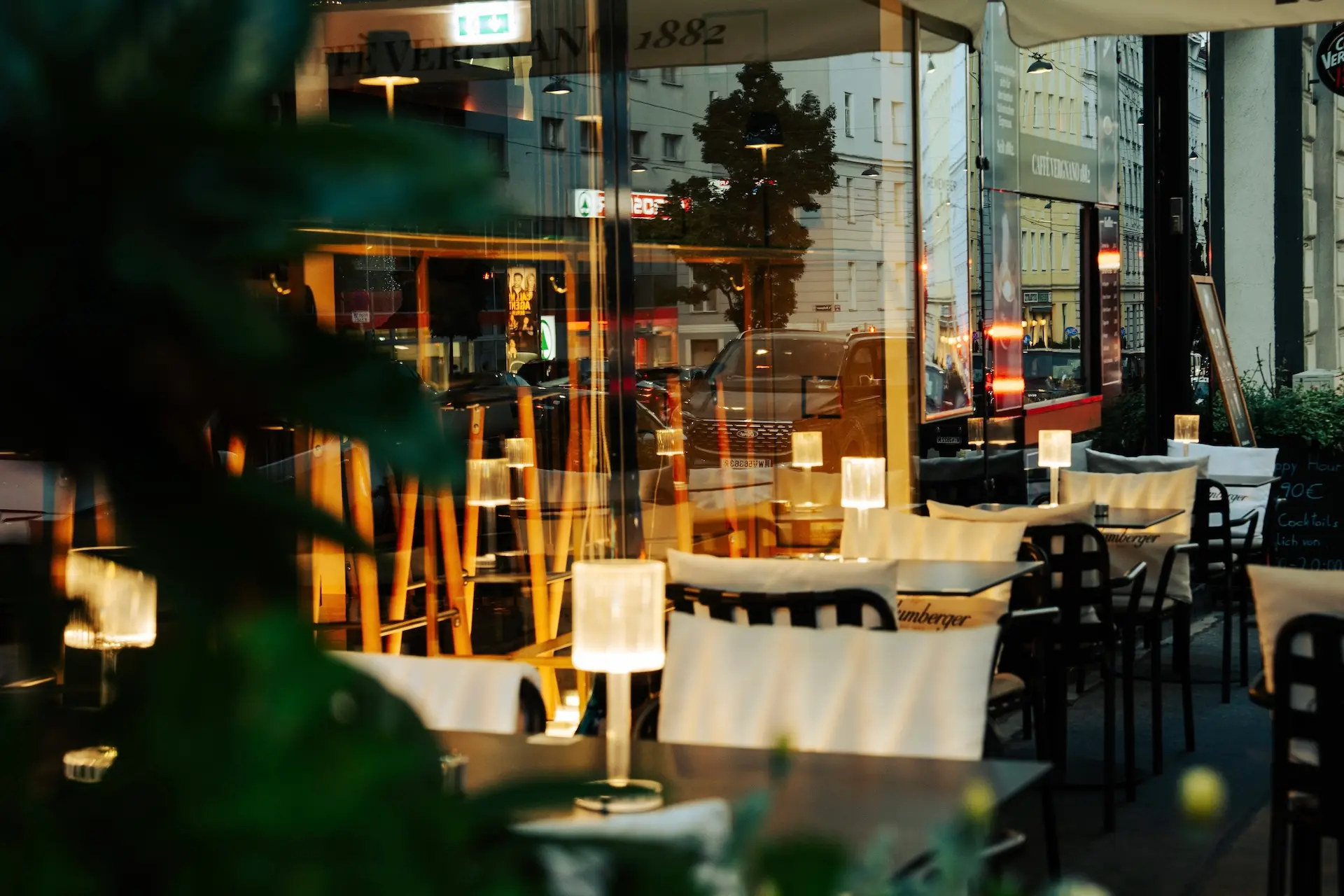 Außensitzbereich eines Cafés mit kleinen Tischen und Stühlen, jeder Tisch von einer kleinen Lampe beleuchtet. In den großen Fenstern spiegeln sich die Gebäude der Stadt und die Straßenbeleuchtung. Die Szene wirkt am Abend gemütlich und einladend.