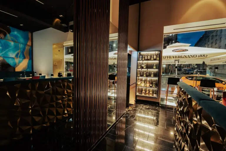 Modernes, stilvolles Café-Interieur mit geometrischen goldenen Paneelen, reflektierendem schwarzem Bodenbelag und einer Wand mit beleuchteten Kaffeeprodukten. Große Fenster zeigen eine Straßenszene mit einem Regenschirm des Caffè Vergnano 1882 und einem gelben Taxi davor.