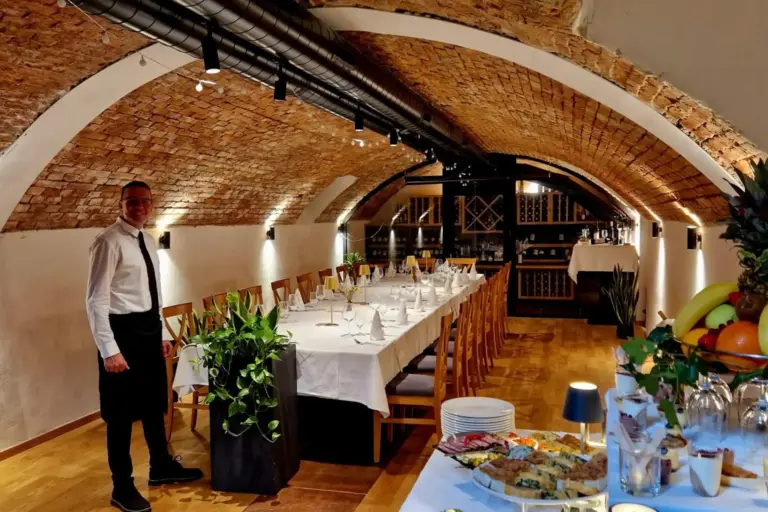 Ein Kellner steht neben einem langen, elegant gedeckten Esstisch unter einer gewölbten Ziegeldecke in einem gemütlichen, warm beleuchteten Restaurant. Im Vordergrund ist ein Buffettisch mit Lebensmitteln und Obst zu sehen.