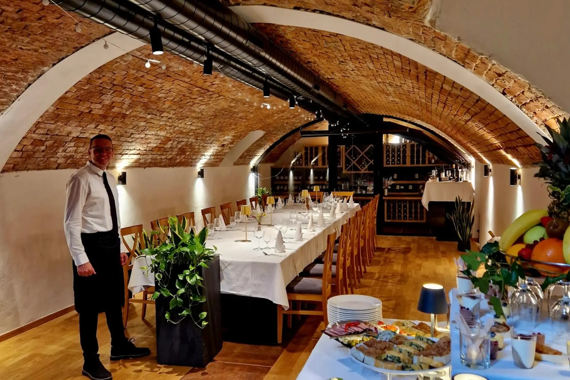 Ein Kellner steht neben einem langen, elegant gedeckten Esstisch unter einer gewölbten Ziegeldecke in einem gemütlichen, warm beleuchteten Restaurant. Im Vordergrund ist ein Buffettisch mit Lebensmitteln und Obst zu sehen.