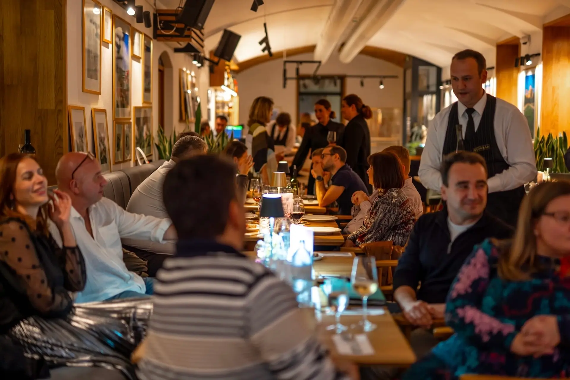Eine lebhafte Restaurantszene mit Menschen, die an Tischen sitzen, sich unterhalten und ihr Essen genießen, während ein Kellner in der Nähe steht. Der Raum ist warm beleuchtet und bietet eine gemütliche, geschäftige Atmosphäre.