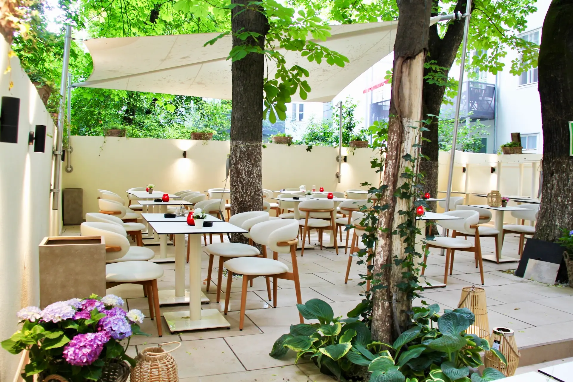 Caféterrasse mit weißen Tischen und Stühlen unter großen Bäumen und einem weißen Vordach. Topfpflanzen und Blumen schmücken den gefliesten Boden und schaffen eine gemütliche und einladende Atmosphäre.